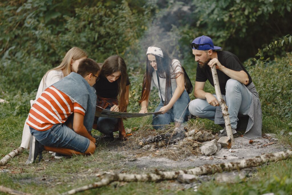 A diverse group of young adults gather around a campfire, exploring a map during a hiking adventure.